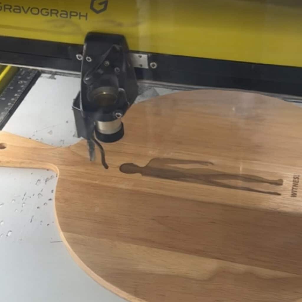 A Gravograph laser engraver customizing a wooden pizza peel with an alien-like design. The laser head is active on the light brown wood.