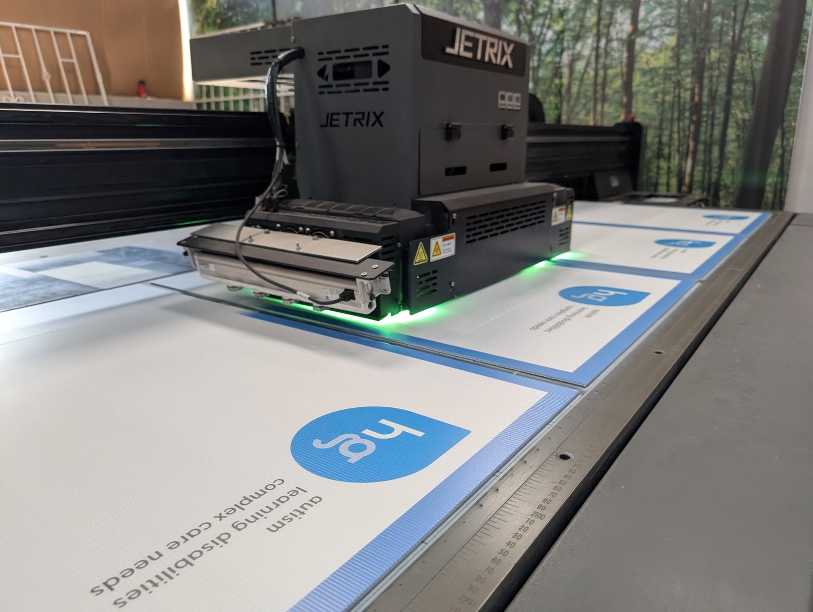 JETRIX UV flatbed printer producing custom corrugated plastic signs with blue 'hg' logo & text. Green UV light illuminates print.