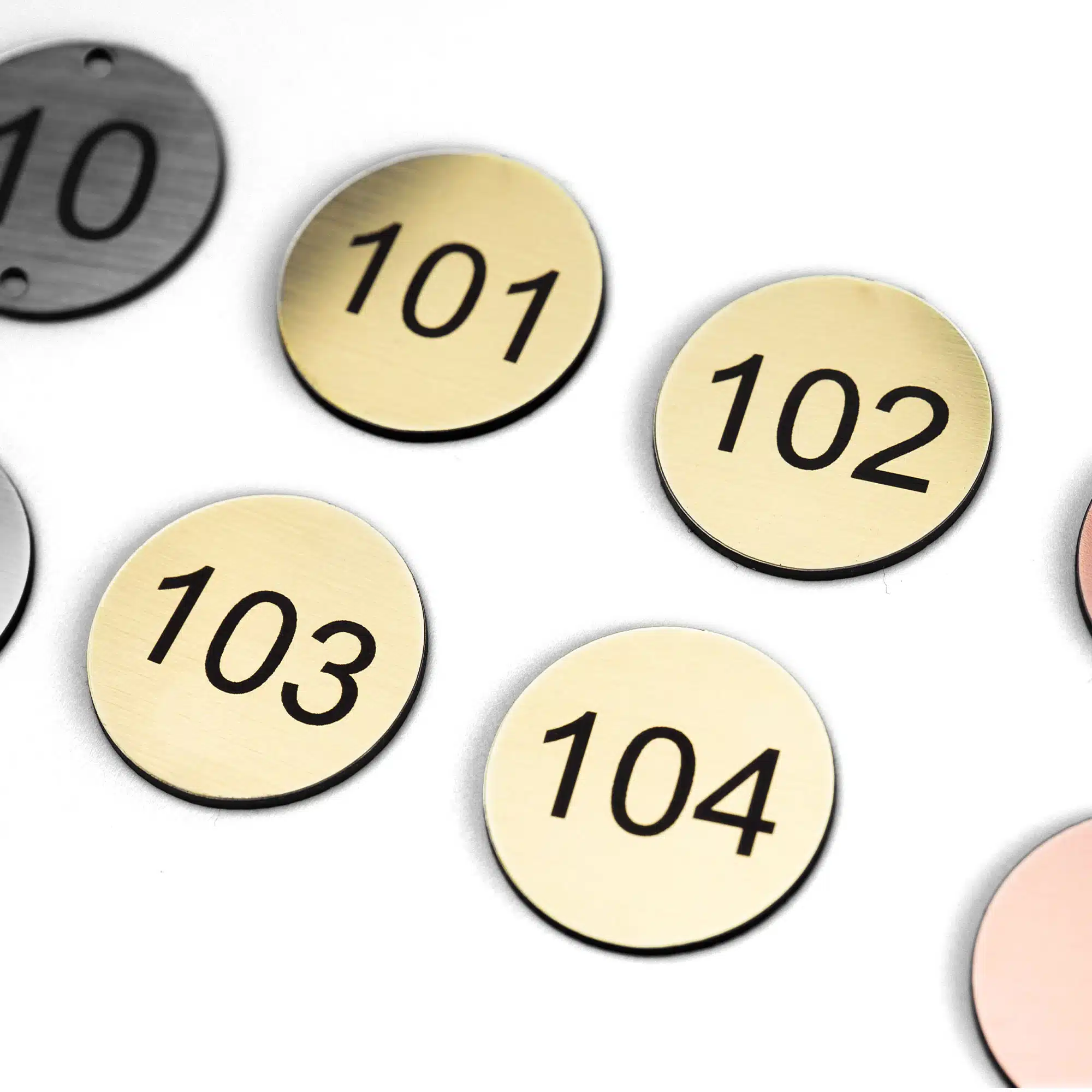Assorted numbered metal tags including brushed gold (101-104), grey (10), and copper discs for tables, rooms or lockers.