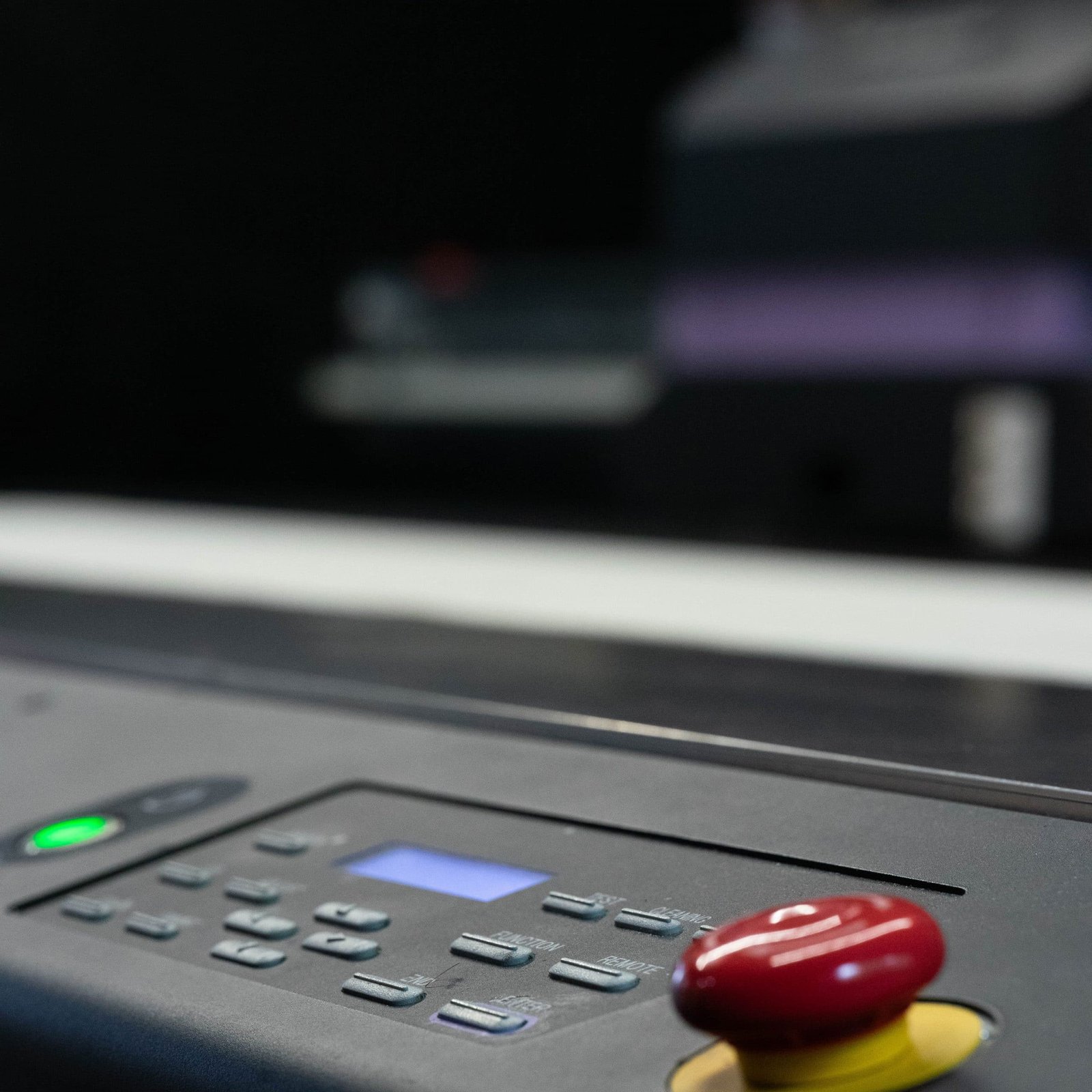 Close-up of a dark grey industrial control panel featuring a red emergency stop button, glowing green light, and keypad.
