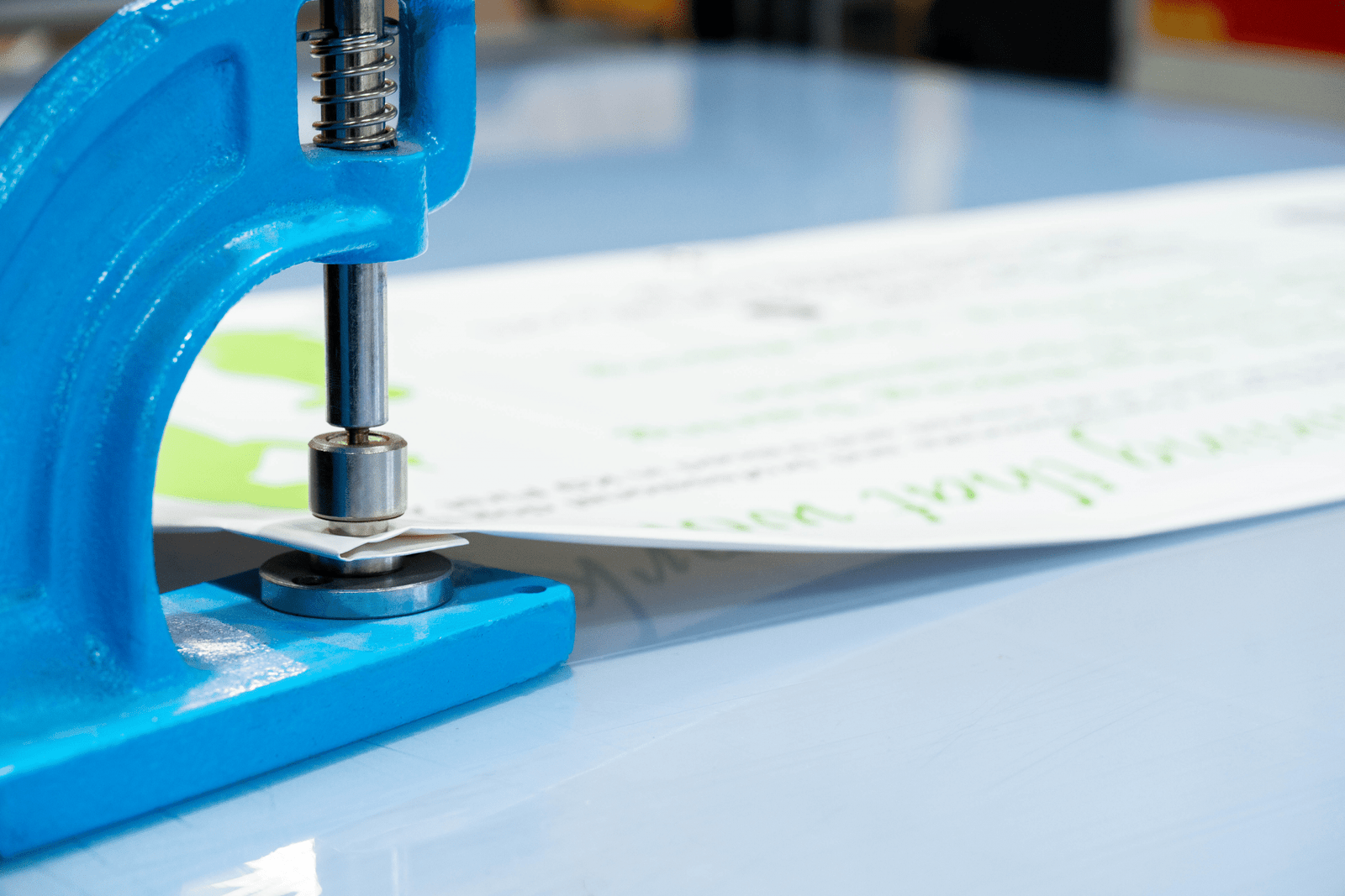A close-up of a bright blue manual eyelet press machine securing a white document with green text on a light blue table.