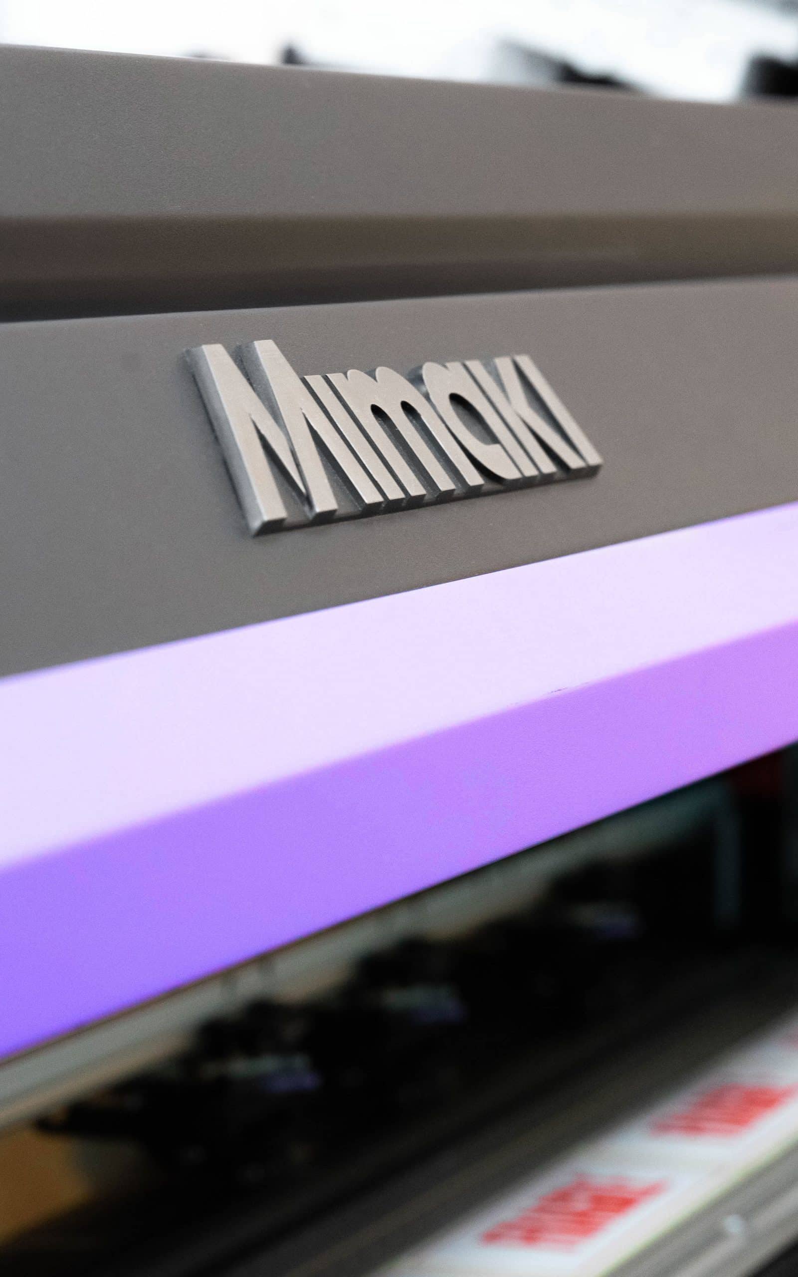 Close-up of a Mimaki industrial printer, featuring its metallic logo, a glowing purple operational light, and blurred print media.