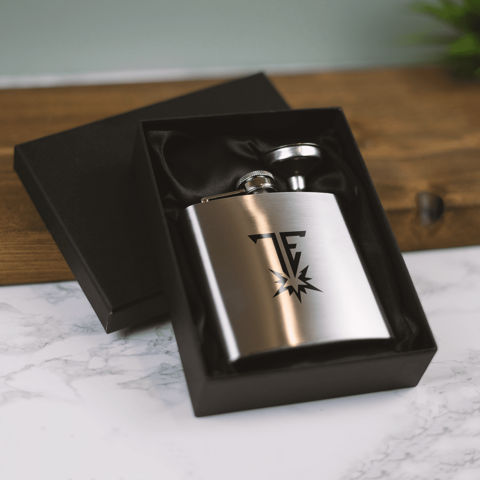 Brushed stainless steel hip flask with custom engraved 'TF' and starburst logo resting in a black satin-lined gift box on a marble surface.