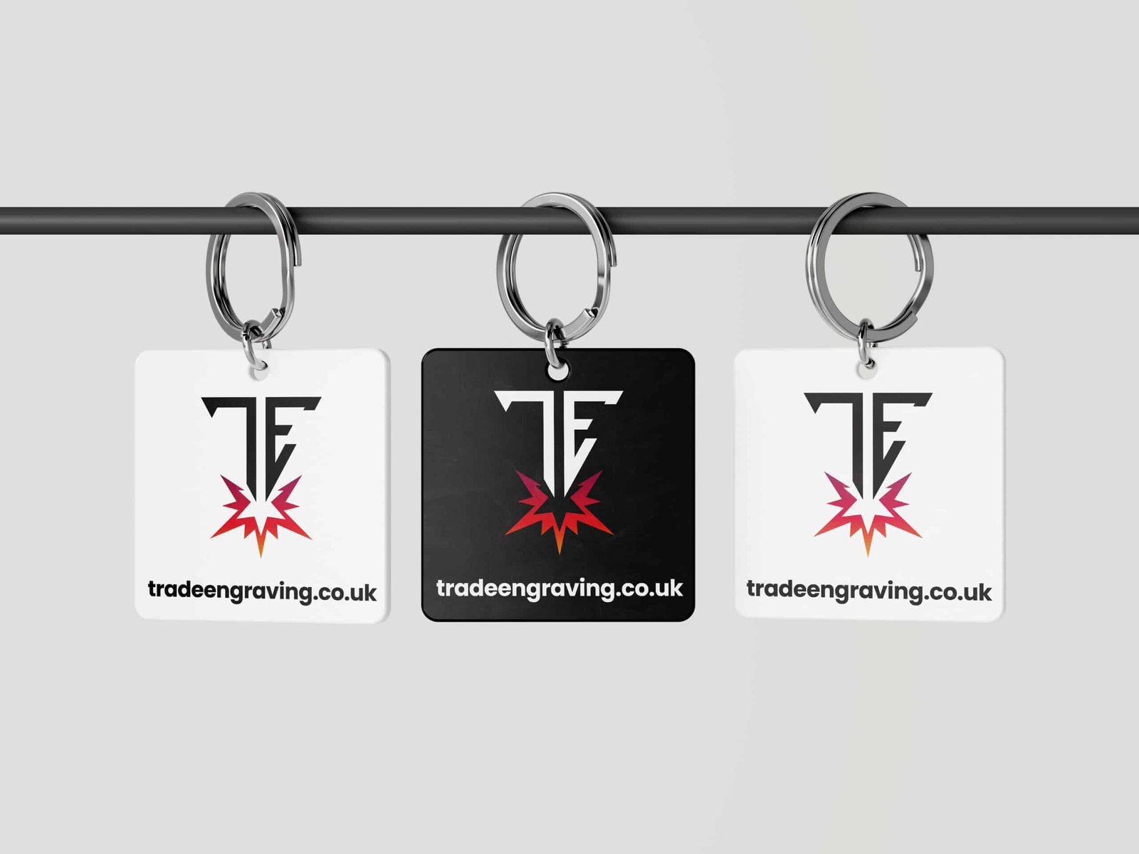 Three keychains displaying the Trade Engraving logo and website, hanging from a bar. Two are white, one is black.