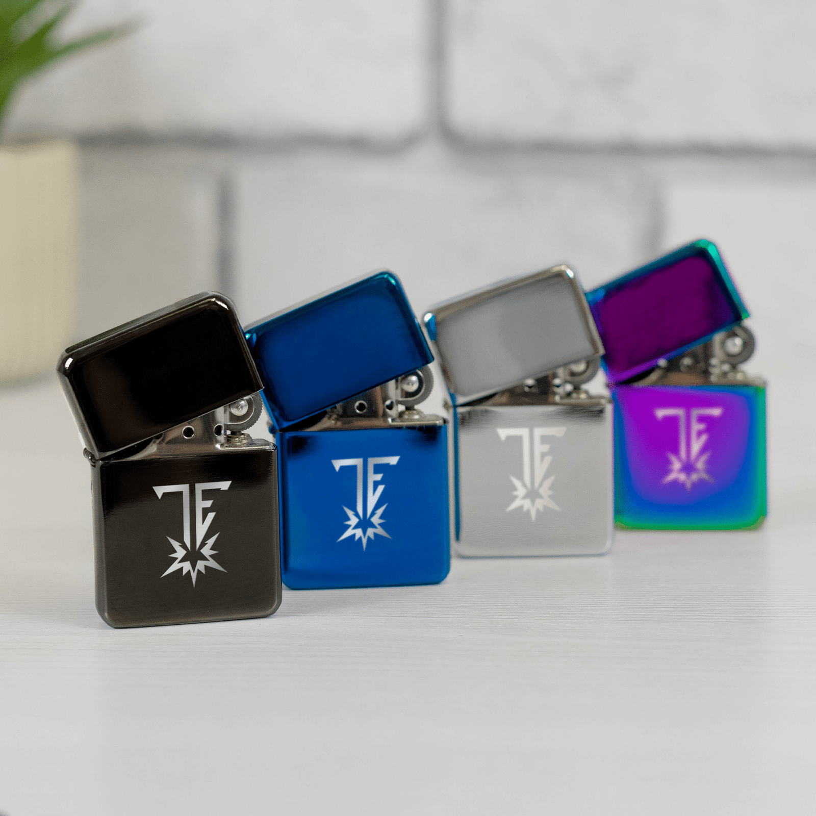 Four sleek lighters in black, blue, silver, and iridescent rainbow colors, each engraved with a unique stylized logo, displayed on a white surface.