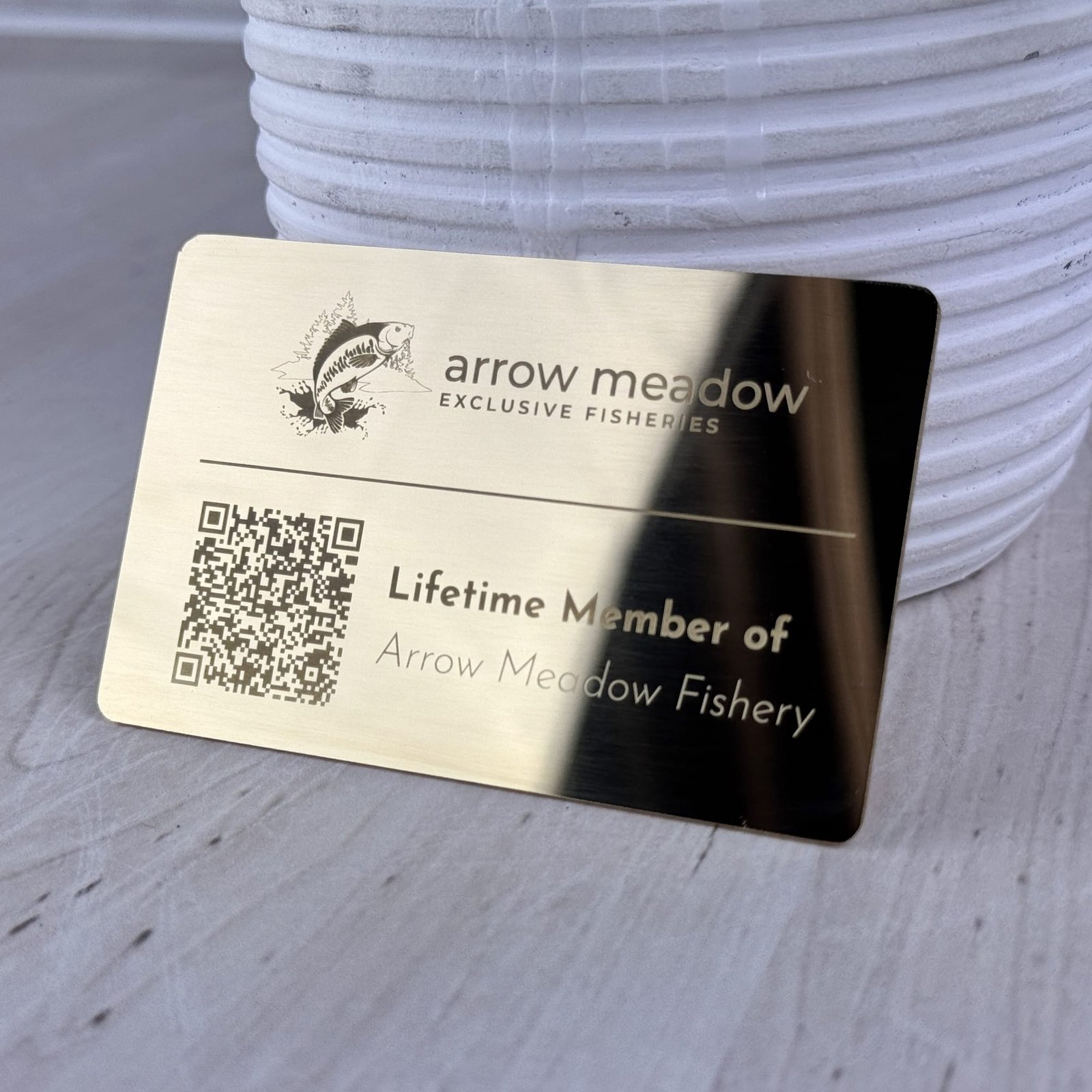 A shiny golden metallic lifetime membership card for Arrow Meadow Fishery, featuring a jumping fish logo and QR code.