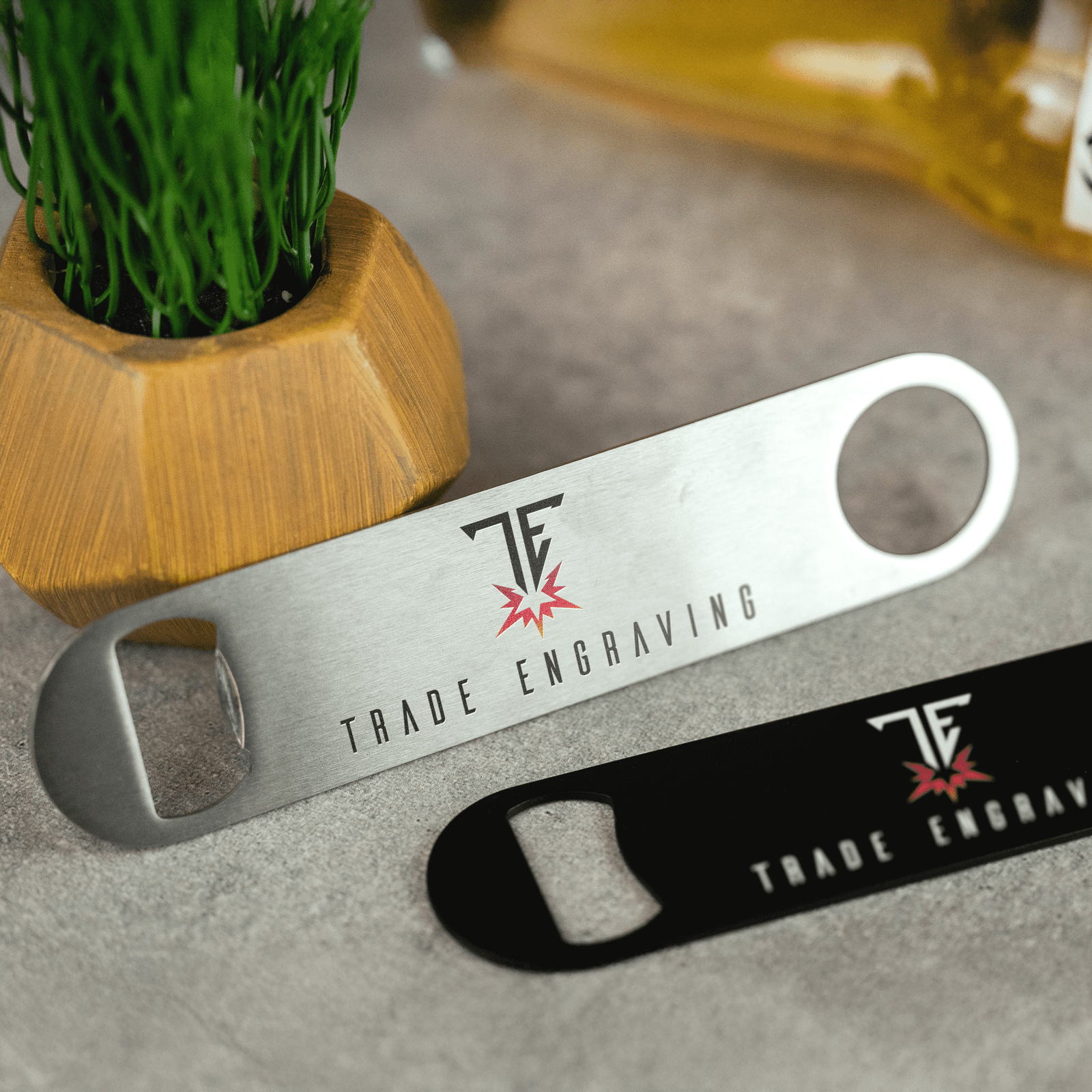 Two custom-engraved metal bar blade bottle openers, one silver and one black, with 'Trade Engraving' logo on a grey surface.