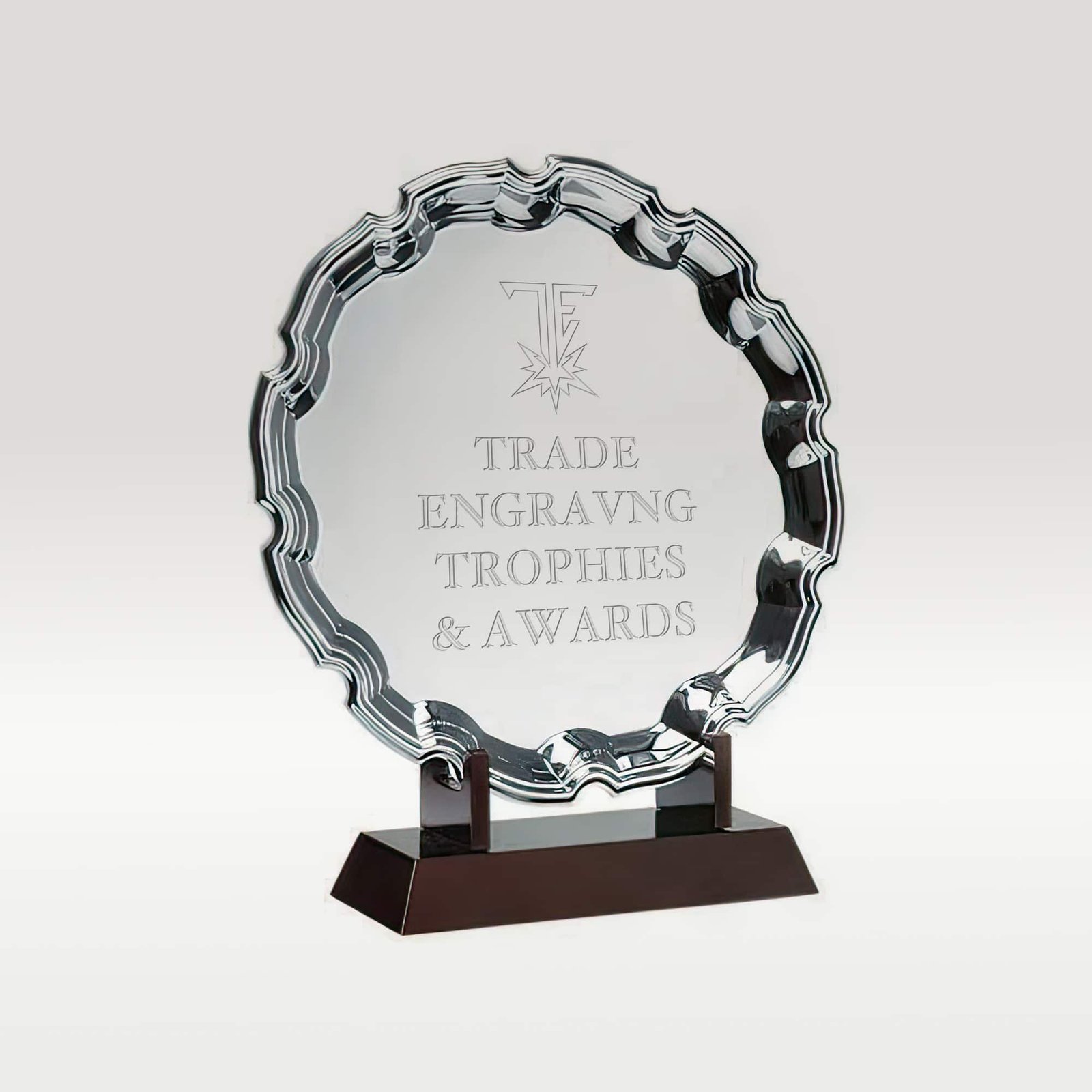 Circular silver award plaque with an ornate wavy edge on a dark brown base, engraved with 'TRADE ENGRAVNG TROPHIES & AWARDS'.