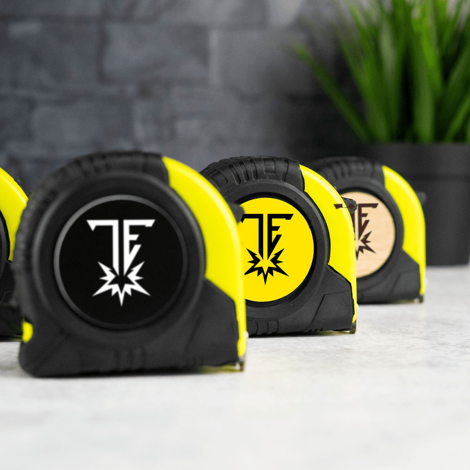 Row of yellow and black branded tape measures on a light surface with a brick wall and green plant background.