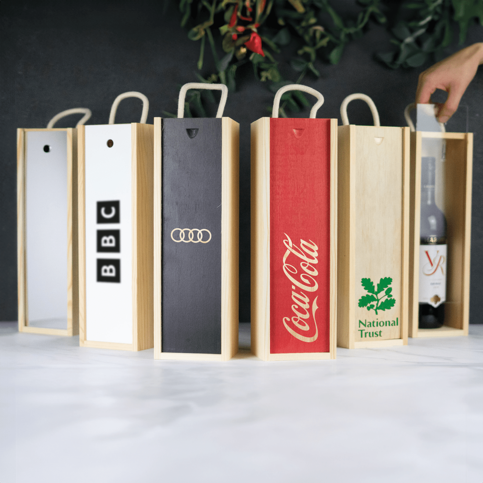 Row of custom branded wooden gift boxes featuring BBC, Audi, Coca-Cola, and National Trust logos, with a hand placing a wine bottle.